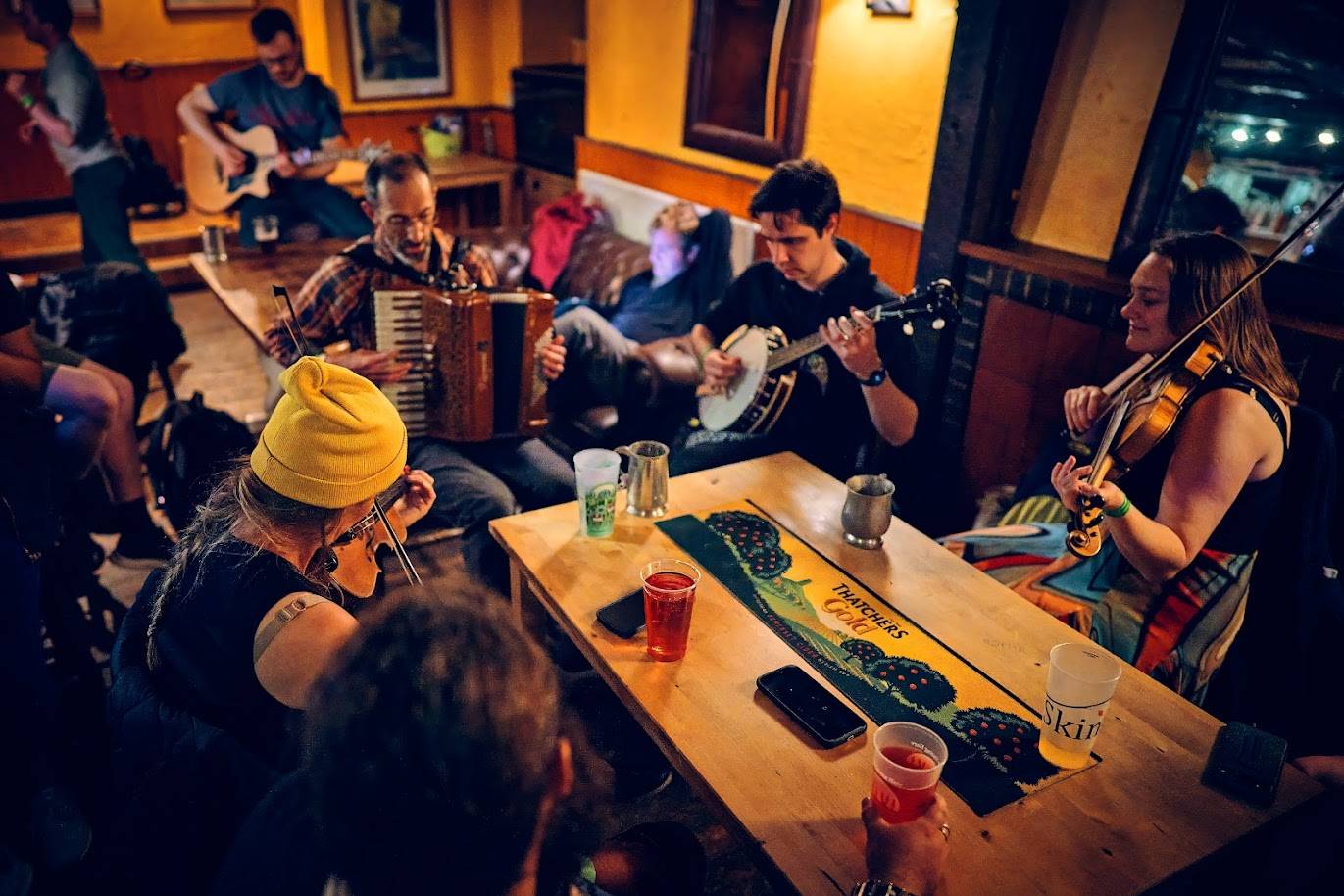 A group of musicians sit around a pub table enjoying a music session. Instruments played include fiddle, accordion, guitar and banjo